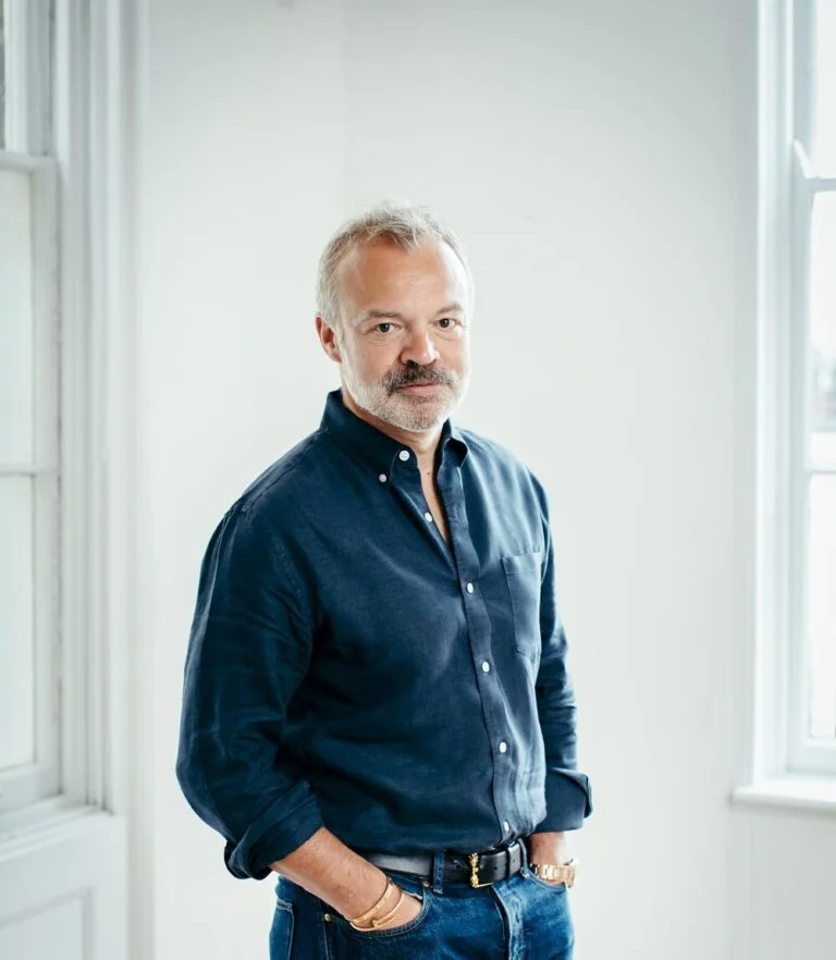 Graham Norton, author and talkshow host, standing in a white room wearing dark blue clothing